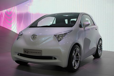 Pearl-white small hatchback concept with sharp headlamps and an iQ Concept plate, in front of a glowing pink and white LED wall.