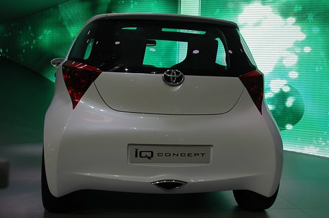 Rear of a pearl-white rounded hatchback concept with red tail lamps and an iQ Concept plate, against a green LED wall. Opens a larger view