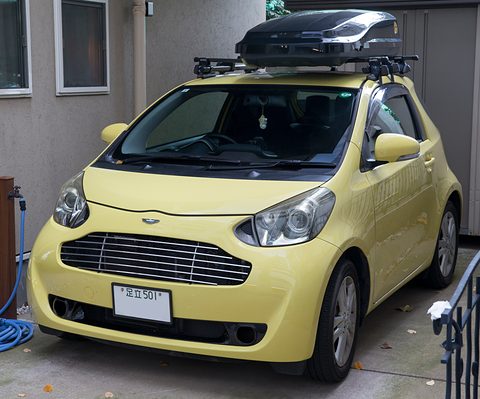 Yellow city car with a slatted Aston-style grille and winged badge, a black roof box on top, parked by a wall. Opens a larger view