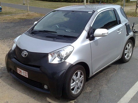 Silver city car with black-taped front and side panels, a windscreen sensor and odd mirrors, parked on gravel. Opens a larger view