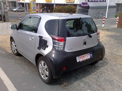 Rear of a silver city car with black tape over its lower body, parked on a gravel verge by a street. Opens a larger view