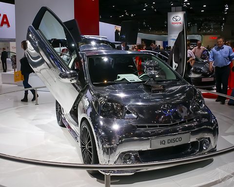 City car covered in small mirror tiles with scissor doors raised, on a turntable among show visitors. Opens a larger view