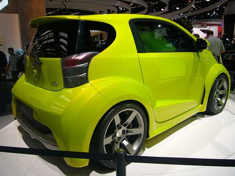 Rear of a lime-green city car with a roof spoiler, flared arches and large wheels behind a rope barrier, visitors around. Opens a larger view