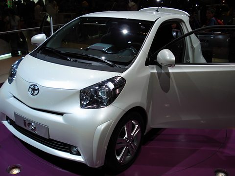 Pearl-white small hatchback with its driver’s door open on a purple-lit display floor. Opens a larger view