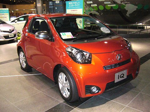 Bright orange city car with a front spoiler and fog lamps on a polished showroom floor. Opens a larger view