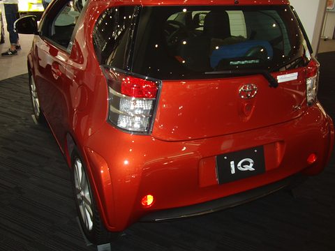 Rear of a red city car with a roof spoiler and an iQ plate on a dark showroom floor. Opens a larger view
