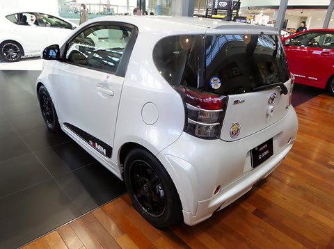 Rear of a white city car with black wheels, GRMN stripes and a Gazoo Racing sticker on a wooden floor. Opens a larger view