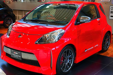 Orange city car with a deep front bumper, large alloy wheels and red brake calipers on a showroom floor. Opens a larger view