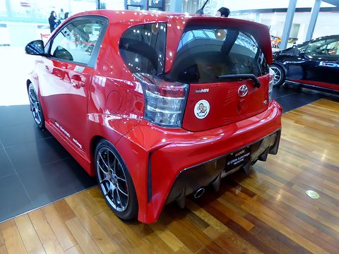 Rear of a red city car with a roof spoiler, rear diffuser and large wheels on a wooden showroom floor. Opens a larger view