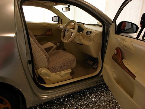 Beige seats, a beige dashboard and a wood-effect door panel seen through the open driver’s door of a small car on gravel. Opens a larger view