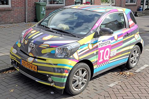 City car wrapped in bright stripes of pink, yellow and blue with a large iQ panel, parked on brick paving. Opens a larger view