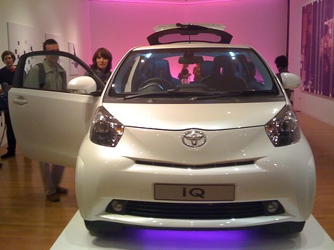 Pearl-white city car with its doors open on a white plinth under pink light, visitors at the door. Opens a larger view