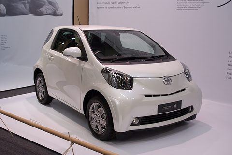 White city car on a white display plinth with text panels on the wall behind. Opens a larger view