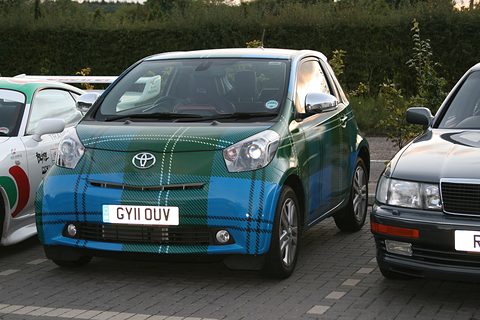 City car wrapped in blue and green tartan, parked on block paving between a white car and a dark saloon. Opens a larger view