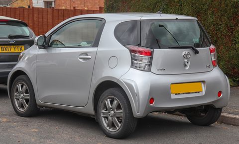 Rear of a silver short hatchback with tall tail lamps and a blanked yellow plate, parked by a fence and hedge. Opens a larger view