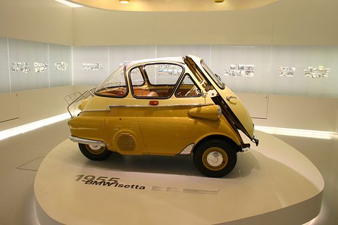 Yellow bubble car with its whole front swung open, showing the steering wheel and bench inside. Opens a larger view