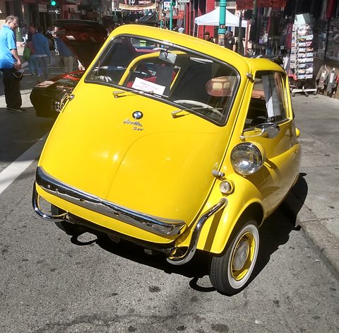 Yellow bubble car seen from the front on a street, stalls and visitors behind. Opens a larger view