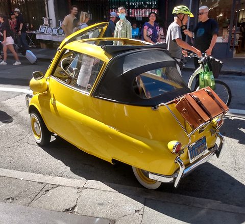 Yellow bubble car with a black folded roof and a leather case on the rack, people around. Opens a larger view