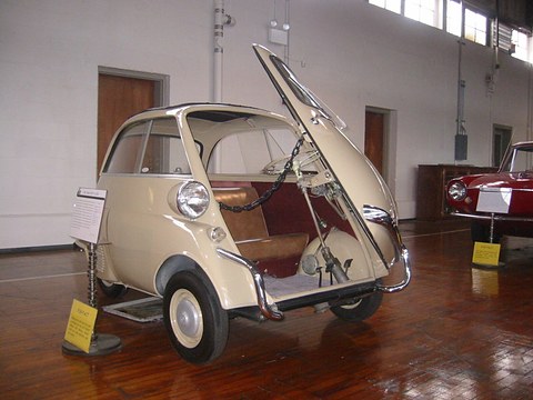 Cream bubble car with its front door open on a wooden museum floor. Opens a larger view