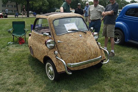 Bubble car whose whole body is clad in woven wicker, parked on grass with people behind. Opens a larger view