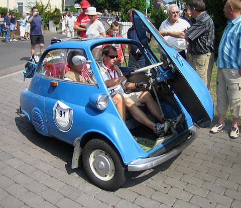 Blue bubble car with rally number 91 and its front door open, the driver seated, onlookers around. Opens a larger view