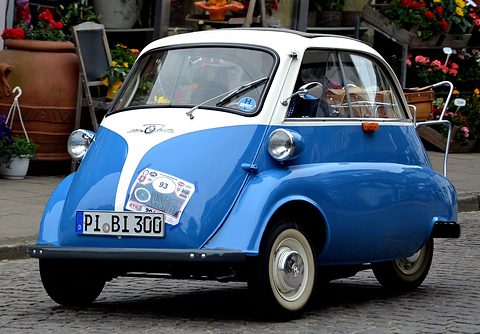 Blue and white bubble car with rally plates parked in front of a flower stall. Opens a larger view