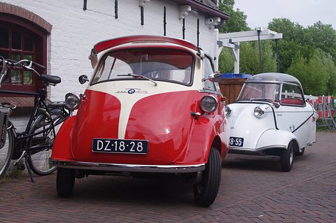 Red and white bubble car head-on next to a white Messerschmitt, bicycles at left. Opens a larger view