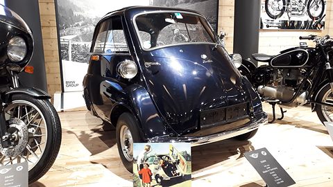 Dark blue bubble car between two BMW motorcycles on a wooden museum floor. Opens a larger view