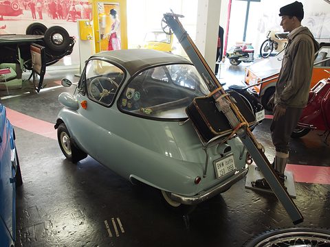 Pale green bubble car from behind with a wooden ladder and suitcase strapped on, a mannequin beside. Opens a larger view