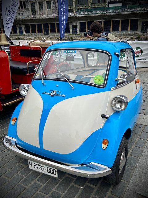 Blue and white bubble car on cobbles seen from the front left, other cars behind. Opens a larger view