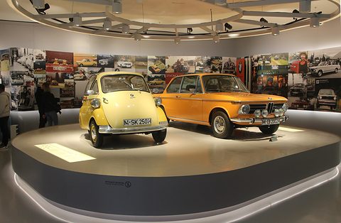 Yellow bubble car next to an orange saloon on a round display under a wall of period photographs. Opens a larger view