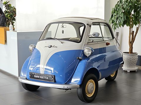 Blue and white bubble car on a grey tiled floor beside a potted plant, seen from the front left. Opens a larger view