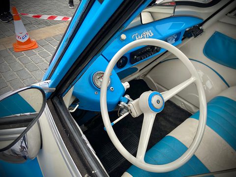White two-spoke steering wheel and a round speedometer against blue painted panels. Opens a larger view