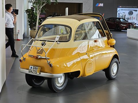 Yellow bubble car seen from behind on a tiled floor with a chrome rack, a man standing behind. Opens a larger view