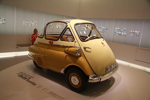 Yellow bubble car with a cream roof on a round white plinth in a softly lit museum room. Opens a larger view