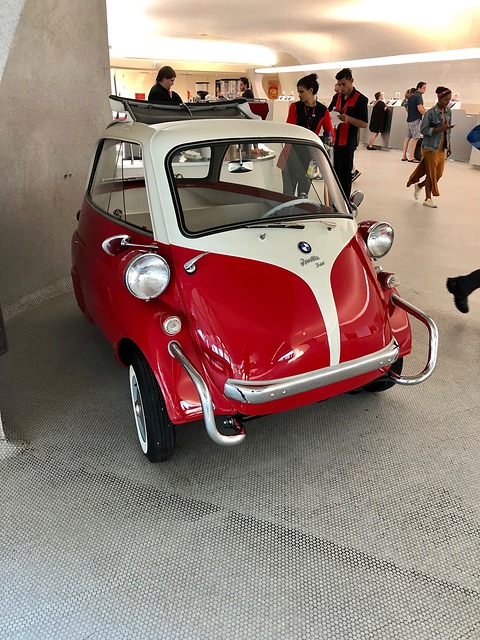 Red and cream bubble car seen from the front in a white terminal, visitors walking behind. Opens a larger view