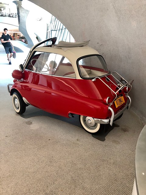 Red and cream bubble car with a luggage rack in a curving white terminal hall. Opens a larger view