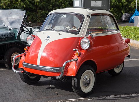 Coral and ivory bubble car with tubular bumpers parked on tarmac, seen from the front right. Opens a larger view