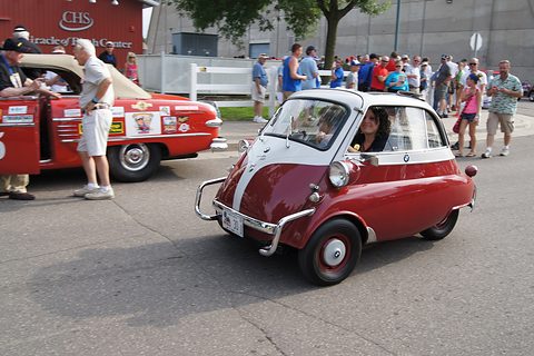 Maroon and white bubble car driving past a crowd at a show, the driver visible. Opens a larger view