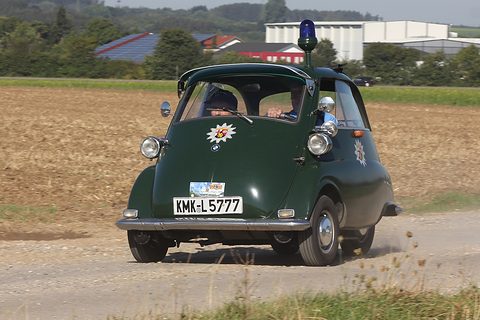 Dark green bubble car with a blue roof beacon and edelweiss emblem driving on a farm track. Opens a larger view