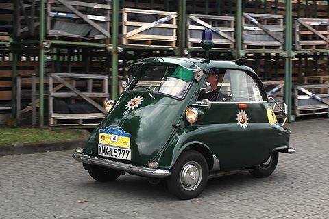 Dark green bubble car with a blue roof light and rally plate, stacked crates behind. Opens a larger view