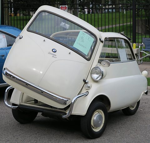 Ivory bubble car with large bumpers and sealed-beam lamps, seen from the front left by a fence. Opens a larger view