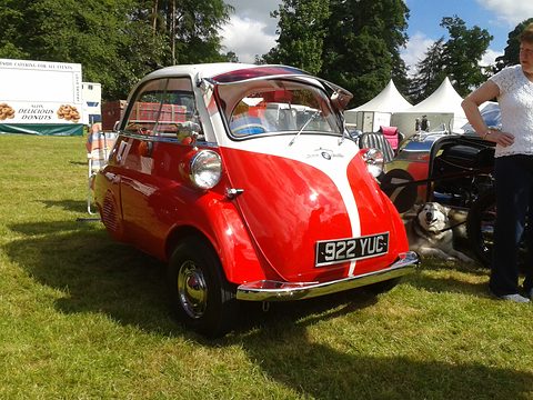 Red and white bubble car on a lawn with a British plate, trees and marquees behind. Opens a larger view