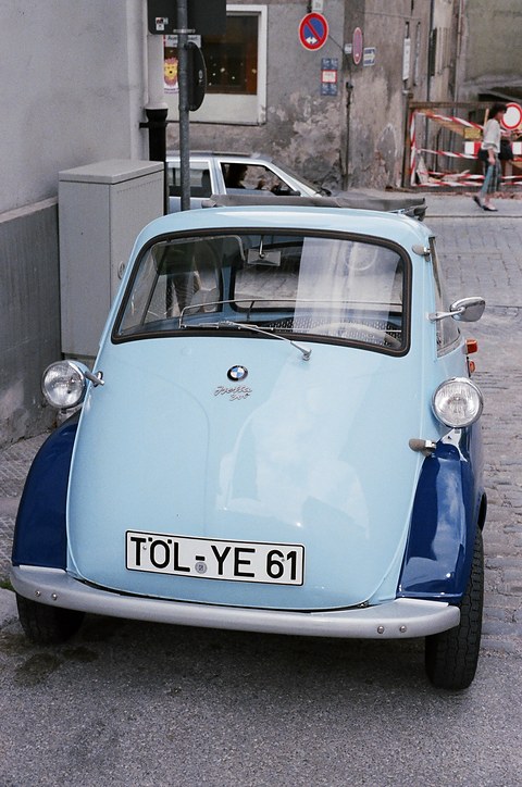 Pale and dark blue bubble car seen straight from the front in a narrow cobbled street. Opens a larger view
