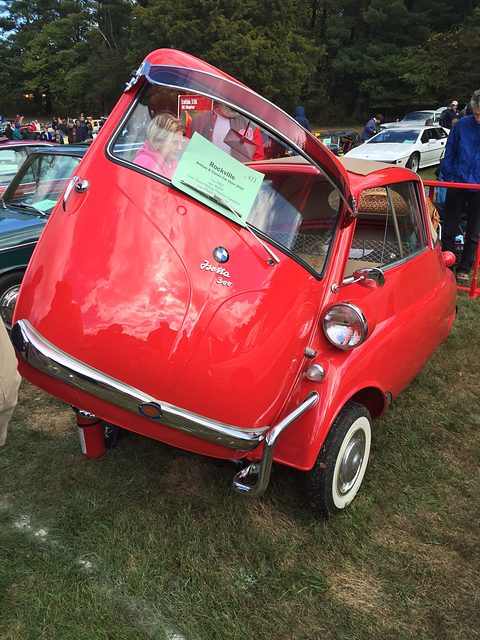 Red bubble car with a fabric roof and door open on grass, visitors behind. Opens a larger view