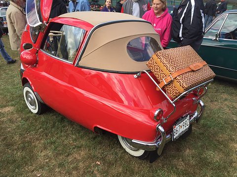 Red bubble car with a tan folding roof and a suitcase on its rack, on grass among people. Opens a larger view