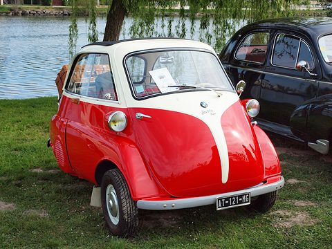 Red and white bubble car on grass by a lake, seen from the front left. Opens a larger view