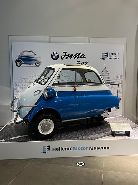 Blue and white bubble car on a small plinth in front of a museum backdrop with the Isetta name. Opens a larger view