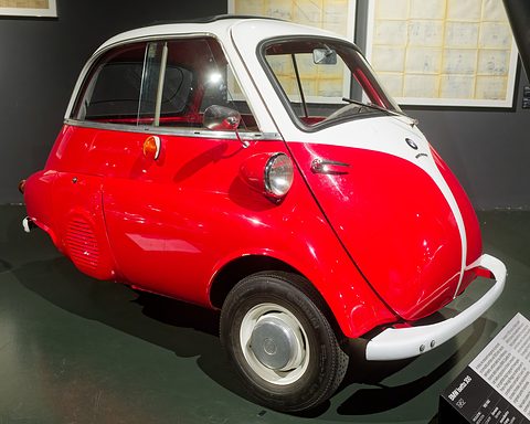 Red and white bubble car with whitewall tyres on a dark museum floor beside a caption board. Opens a larger view