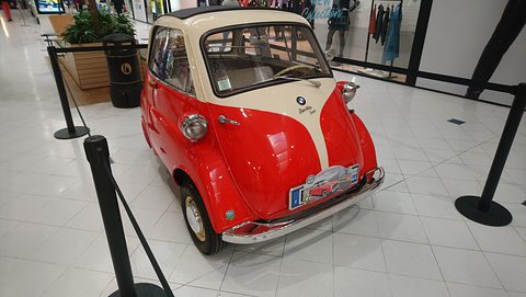 Red and cream bubble car with an event plaque on its bumper, behind rope posts on white tiles. Opens a larger view